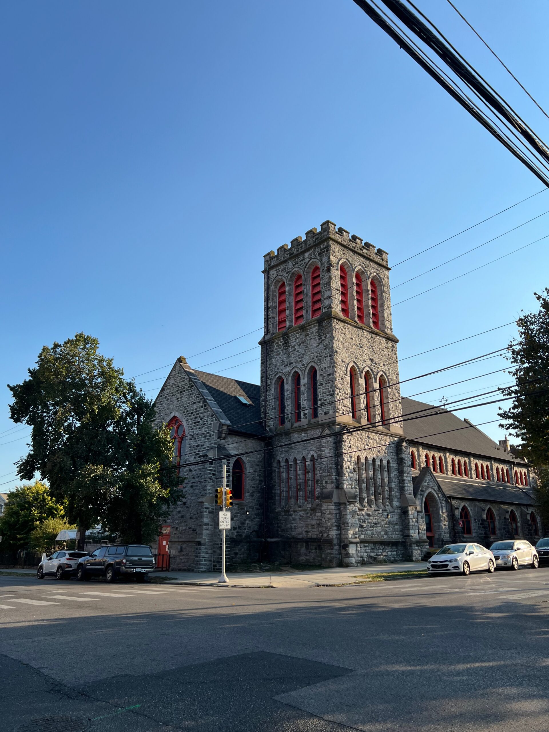 The school building seen from the far corner of 47th Street and Kingsessing Avenue on a sunny day. The building is an architecturally dramatic former church, with a three-story corner tower rising to a crenellated top, and arched windows with orange details set against stone walls.