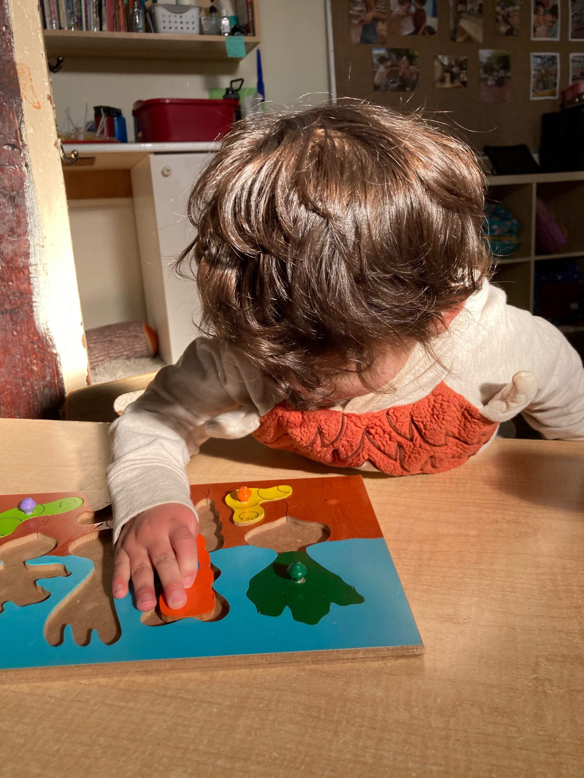 A young child tries fitting a carrot-shaped piece into a wooden vegetable-themed puzzle.